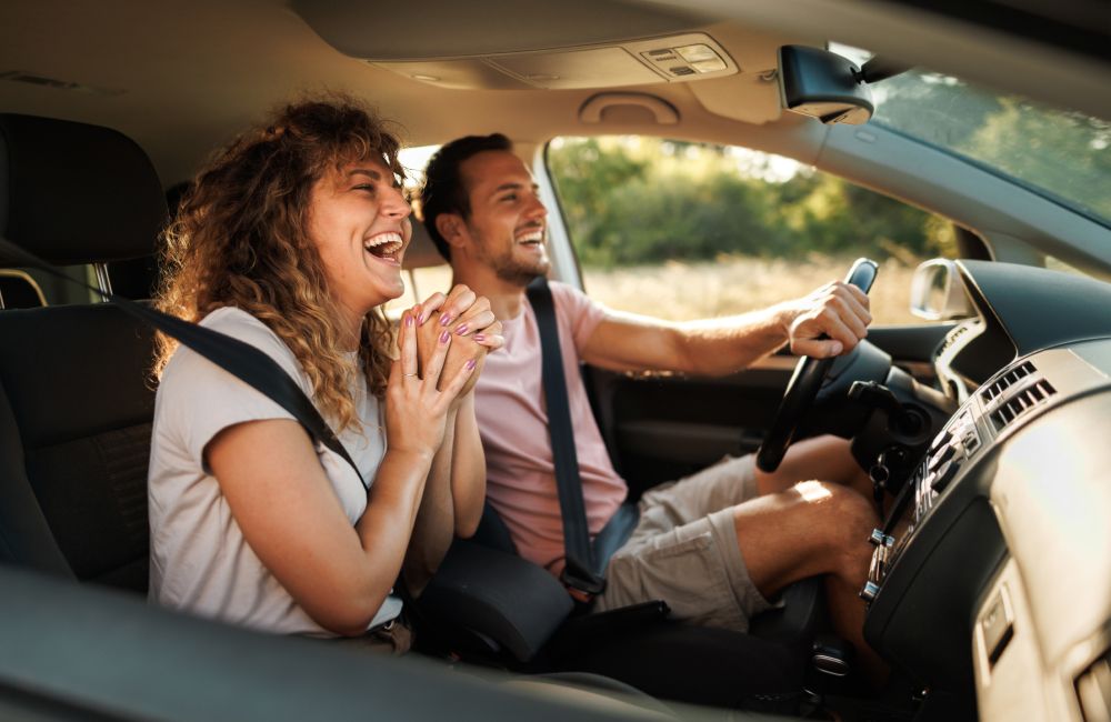Ein heterosexuelles Pärchen sitzt gemeinsam im Auto, hält Händchen und lacht. 