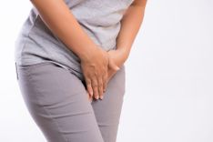 Frau in grauem Shirt und grauer Jeans, die beide Hände über ihren Intimbereich hält